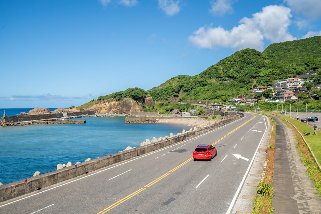 A picturesque view of a coastal road with a red car, blue ocean, and lush green hills on a sunny day.