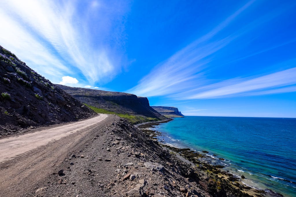 A scenic dirt road winds along a rocky coastline under a vibrant blue sky.