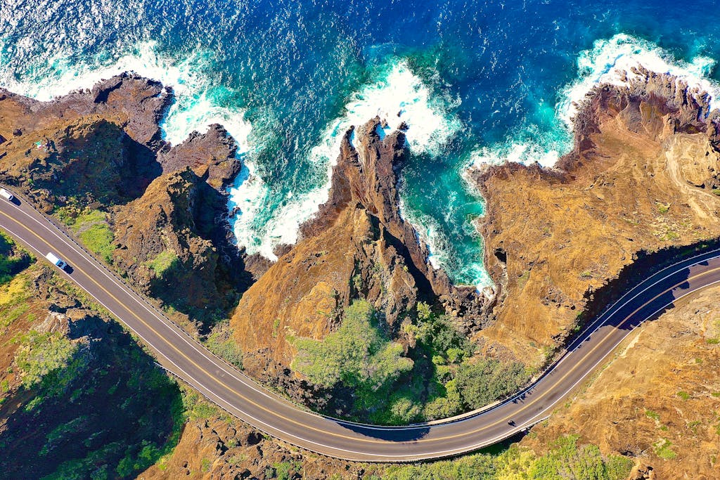 Aerial shot capturing a winding coastal road along a rugged, rocky shoreline with waves crashing into the cliffs.