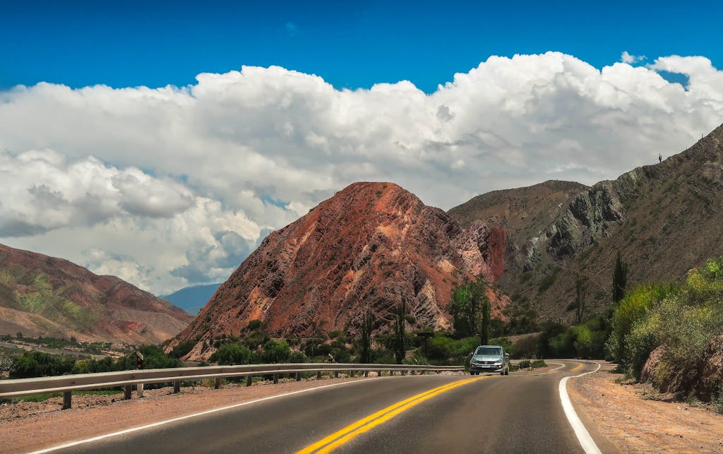 Car journeying on a winding mountain road with a stunning rock formation and vibrant sky.